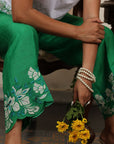 A woman sitting on a chair and wearing white and green co-ord set with flowers in her hand.