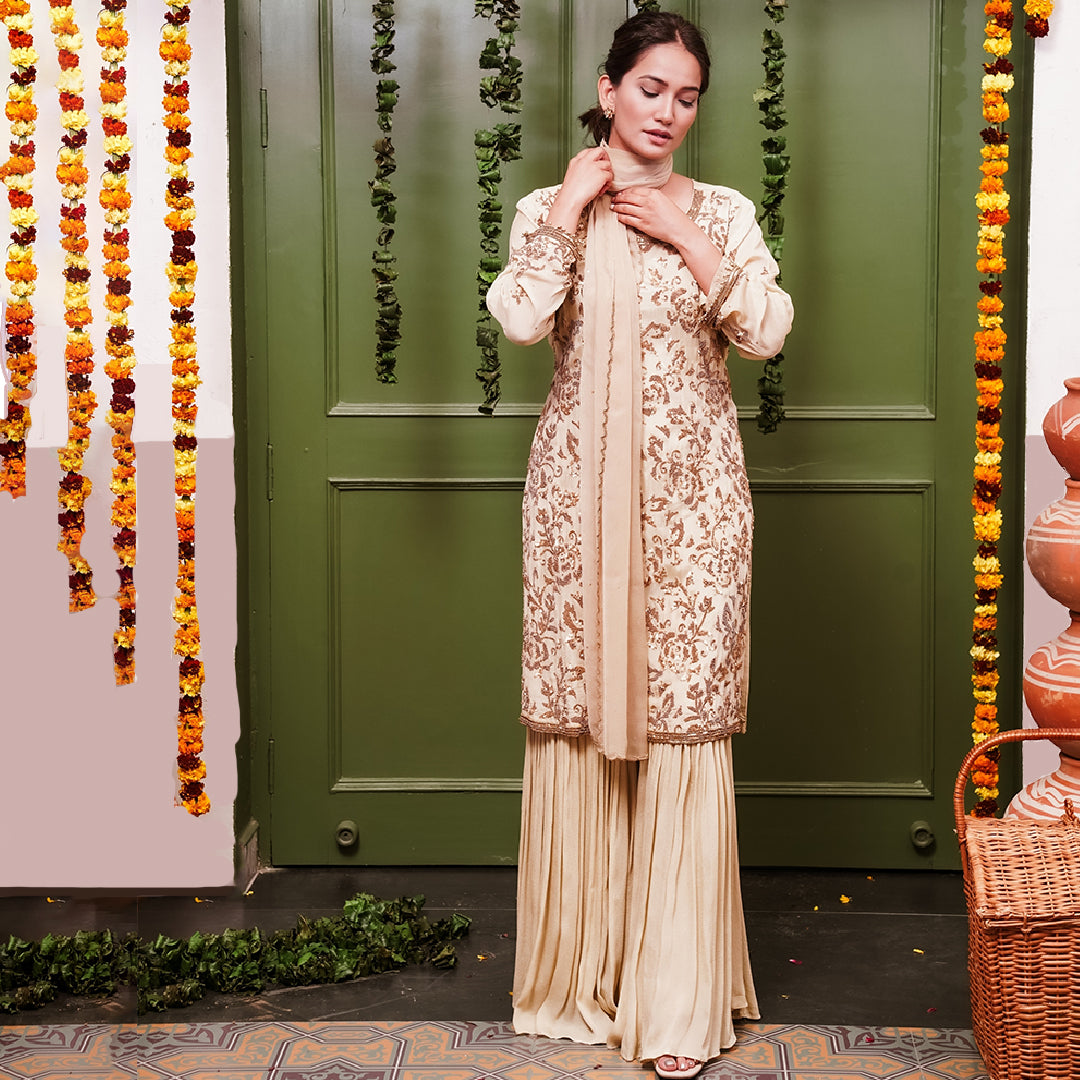 A women wearing beige colored Sharara Suit Set with Dupatta.